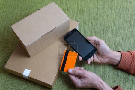 two hands holding mobile phone and orange credit card, symbolizing online shopping, two brown packages on the sofa