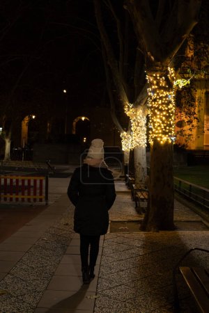 mujer caminando calurosamente sobre su espalda en una noche de invierno con iluminación navideña en los árboles de un parque.
