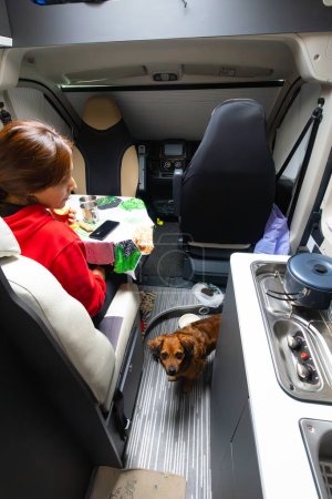 latin american woman inside a camper van having breakfast looking at her dog. you can see swivel seat, kitchen, table, cell phone.