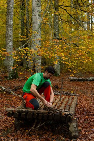 Foto de Un hombre está sentado en un tronco en un bosque. El tronco está hecho de madera y tiene algunas ramas que sobresalen de él. escena pacífica - Imagen libre de derechos