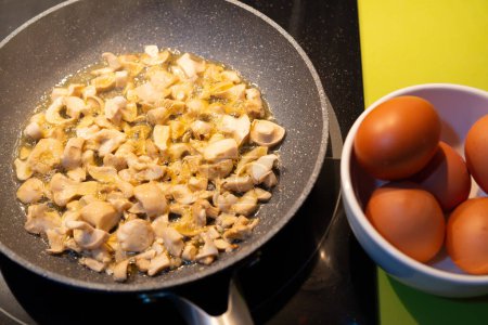 mushrooms cooked in a frying pan with boiling oil on induction hob and green cooking top. next to it fresh eggs in a bowl