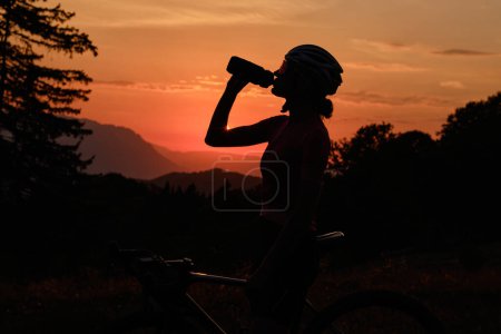 Foto de Silueta de ciclista. Mujer ciclista en ropa de ciclismo y un casco bebe agua de una botella deportiva al atardecer, con una montaña en el fondo - Imagen libre de derechos