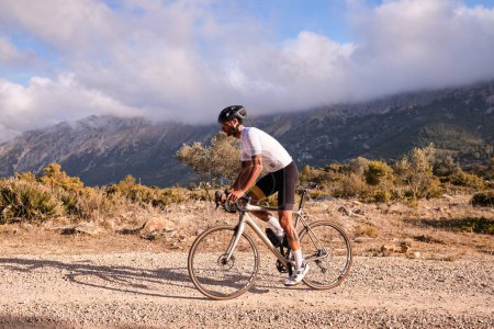 Foto de Ciclista practicando en carretera de grava. Ajuste ciclista masculino montando una bicicleta de grava en una carretera de grava con vistas a las montañas, región de Alicante de España - Imagen libre de derechos