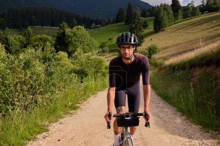 Man cyclist wearing cycling kit and helmet riding gravel bike on gravel road in mountains with scenic view.