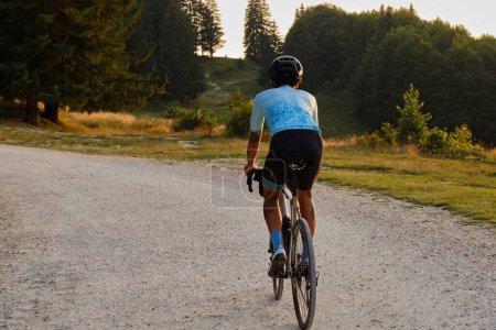 Photo for A fit male cyclist practicing on a gravel road at sunset. He is riding a gravel bike with a view of the mountains. Bucegi Mountains, Romania - Royalty Free Image