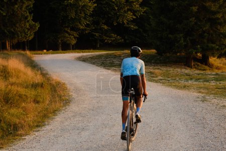 Foto de Hombre ciclista con camiseta de ciclismo azul. Está montando una bicicleta de grava en una carretera de grava al atardecer con vistas a las montañas.Ruta de montaña vacía.Ciclismo aventura de grava en Rumania.Parque Natural de Bucegi - Imagen libre de derechos