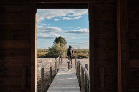 A man is riding over a lake with a background of mountains.Male cyclist is riding a gravel bike on a wooden bridge over a lake.Elche, Alicante, Spain
