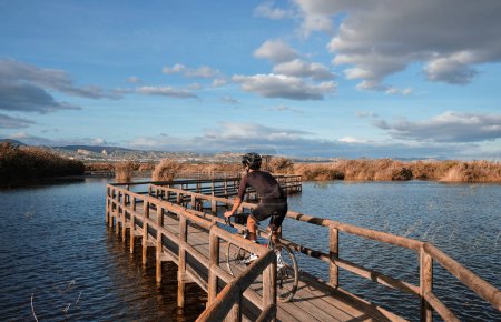 A man is riding over a lake with a background of mountains.Male cyclist is riding a gravel bike on a wooden bridge over a lake.Elche, Alicante, Spain