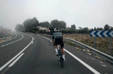 Foto de Ciclista corriendo a través de la niebla en una bicicleta de carretera.Hombre ciclista con kit de ciclismo y casco.Ciclismo en las montañas españolas.Hermosa imagen de motivación de un atlete.Benissa, Alacant, España - Imagen libre de derechos
