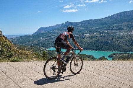 Photo for Man cyclist in helmet and cycling kit riding through mountainous roads. Cyclist on gravel bike with scenic views of turquoise lake in Spain.Gravel riding through the mountains.Guadalest Reservoir. - Royalty Free Image