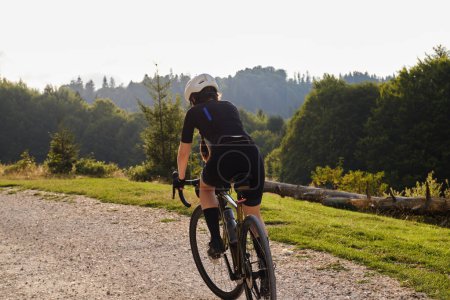 Photo for Woman cyclist wearing black cycling kit. She is riding a gravel bike on a gravel road at sunset with a view of the mountains.Empty mountain road.Cycling gravel adventure in Romania.Bucegi Natural Park - Royalty Free Image