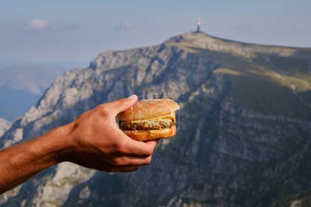 Foto de Savoring the Moment: Close-Up of a Man 's Hand Holding a Juicy Burger with Majestic Mountains in the Background (en inglés). Comida chatarra en las montañas. Momentos de caminata de lujo. Montañas Bucegi, Rumania - Imagen libre de derechos