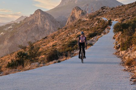 Foto de Fit ciclista mujer está montando una bicicleta eléctrica en una carretera al atardecer con vistas a las montañas y el mar. Camino de montaña vacío. Aventura en bicicleta en España. - Imagen libre de derechos