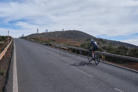 Woman cyclist riding a bicycle on the road with a volcano on the background.Cycling Tenerife.Travels on the road bicycle.Road leading to Mount Teide. Woman cyclist wearing cycling kit and helmet.Spain