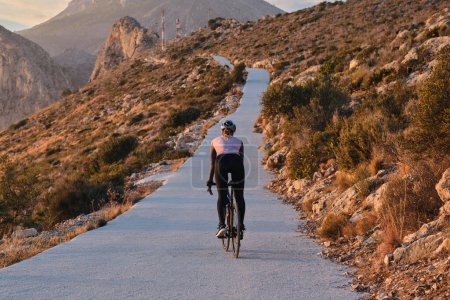 Fit woman cyclist is riding an E-bike on a road at sunset with a view of the mountains and sea. Empty mountain road. Cycling  adventure in Spain.