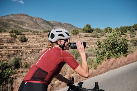 Mujer sedienta ciclista con kit de ciclismo y un casco bebiendo agua de una botella deportiva durante el paseo en bicicleta.