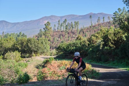 A female cyclist is riding along a scenic mountain gravel route in a forest on Tenerife Island. Cyclist is practicing on gravel road. Beautiful sunny day for cycling. Spain.