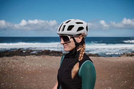 Close up portrait of a female cyclist on Atlantic ocean background. Beautiful woman cyclist is wearing cycling kit, sunglasses and helmet. Sport equipment.Training outdoors. Tenerife, Canary Islands,