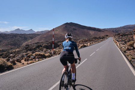 Woman cyclist riding a bicycle on the road with a volcano on the background.Cycling Tenerife.Travels on the road bicycle.Road leading to Mount Teide. Woman cyclist wearing cycling kit and helmet.Spain