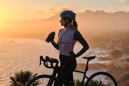 Woman cyclist in cycling kit and a helmet is drinking water from a sports bottle at sunset on mountains background. Cyclist silhouette.