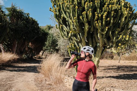 Female cyclist in cycling clothes and a helmet drinks water from a sports bottle during the ride.