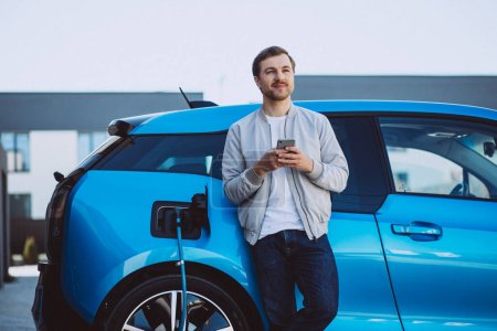 Young man charging electric car at home and using mobile phone
