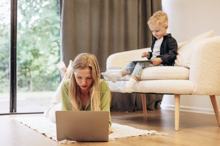 Téléchargez les photos : Femme travaillant sur ordinateur pendant que son fils jouait sur des jeux de tablette - en image libre de droit