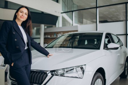 Téléchargez les photos : Femme de vente dans une salle d'exposition de voiture debout par voiture blanche - en image libre de droit