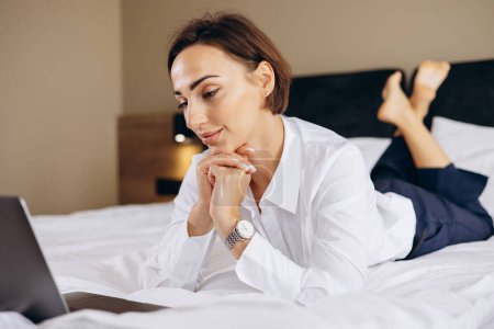 Téléchargez les photos : Femme d'affaires couchée sur le lit et travaillant sur ordinateur dans une chambre d'hôtel - en image libre de droit