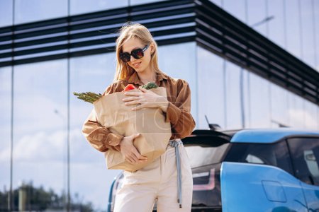 Téléchargez les photos : Femme avec sac en papier d'épicerie par sa voiture électrique - en image libre de droit
