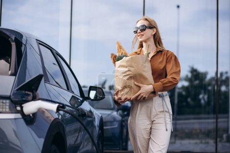 Téléchargez les photos : Femme faisant des courses tandis que la voiture électrique charge - en image libre de droit