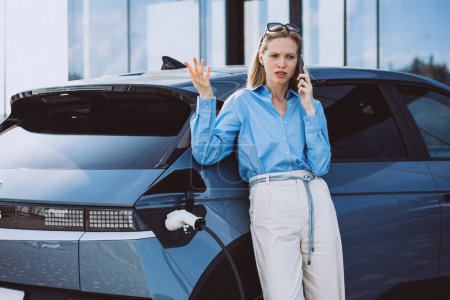 Téléchargez les photos : Femme d'affaires parlant au téléphone et se tenant près de sa voiture électrique - en image libre de droit