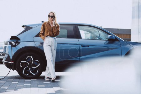 Photo for Woman talking on the phone by her electric car - Royalty Free Image