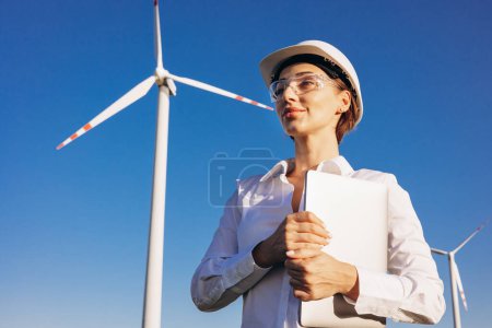 Foto de Arquitecta mujer con portátil con casco blanco y de pie junto a las turbinas del molino de viento - Imagen libre de derechos