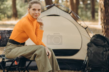 Téléchargez les photos : Femme voyageur assis dans sa chaise près de sa tente dans la forêt - en image libre de droit