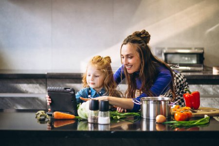 Téléchargez les photos : Mère et fille avec tablette cuisson dans la cuisine - en image libre de droit