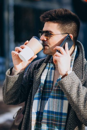 Téléchargez les photos : Jeune homme beau boire du café à l'extérieur et en utilisant le téléphone - en image libre de droit