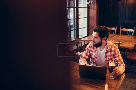 Téléchargez les photos : Youn g homme barbu travaillant sur ordinateur portable dans un bar - en image libre de droit