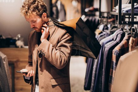 Téléchargez les photos : Jeune homme au magasin de vêtements pour hommes parlant au téléphone - en image libre de droit