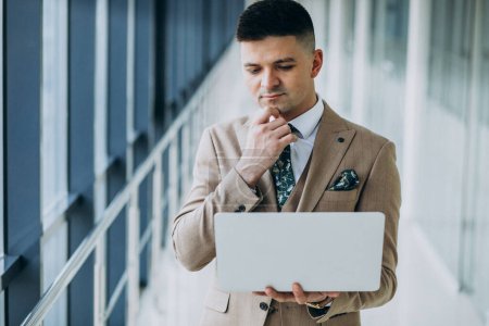 Téléchargez les photos : Jeune homme d'affaires beau debout avec ordinateur portable au bureau - en image libre de droit