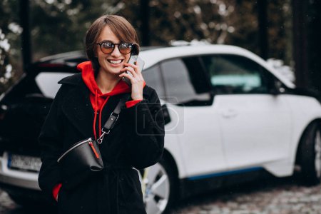 Foto de Mujer cargando electro coche y hablando por teléfono - Imagen libre de derechos
