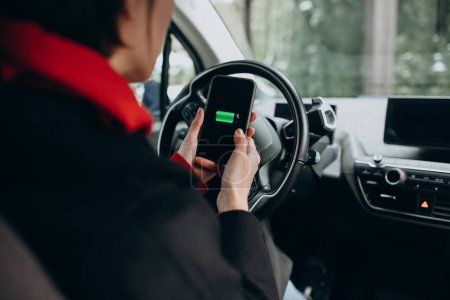 Téléchargez les photos : Femme chargeant sa voiture et regardant le cherger sur son téléphone - en image libre de droit