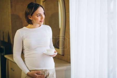 Foto de Mujer embarazada joven de pie junto a la ventana y beber café - Imagen libre de derechos