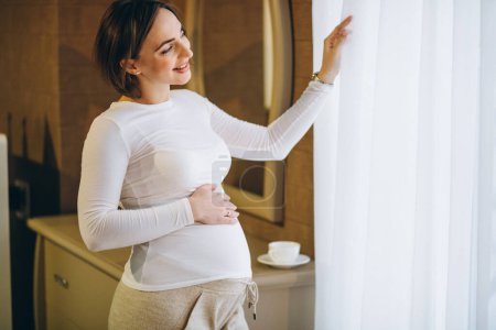 Téléchargez les photos : Jeune femme enceinte debout près de la fenêtre et boire du café - en image libre de droit