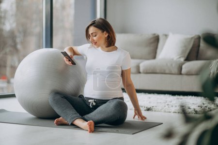 Foto de Young pregnant woman practicing yoga at home and using phone - Imagen libre de derechos