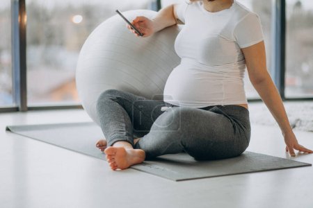 Photo for Young pregnant woman practicing yoga at home and using phone - Royalty Free Image