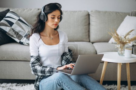 Téléchargez les photos : Jeune belle femme assise à la maison travaillant sur un ordinateur portable dans des écouteurs - en image libre de droit