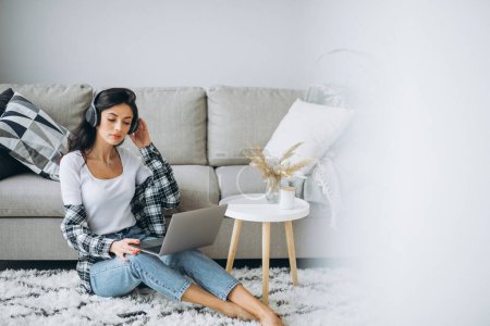 Téléchargez les photos : Jeune belle femme assise à la maison travaillant sur un ordinateur portable dans des écouteurs - en image libre de droit