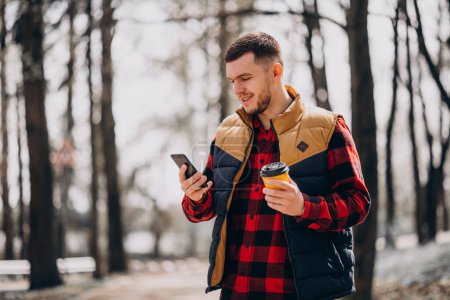 Téléchargez les photos : Jeune homme buvant du café dans le parc et utilisant le téléphone - en image libre de droit