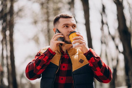Photo for Young man drinking coffee in park and using phone - Royalty Free Image